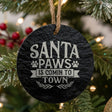 Round black slate Christmas ornament hanging from a jute string on a decorated Christmas tree, laser engraved in white with “Santa Paws is Comin to Town”.