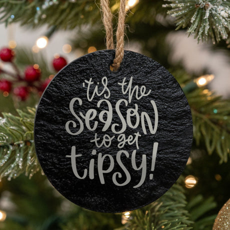 Round black slate Christmas ornament hanging from a jute string on a decorated Christmas tree, laser engraved in white with “Tis the Season to Get Tipsy”.