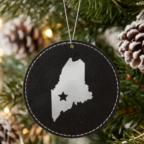 Black leatherette round Christmas ornament laser engraved with the Maine Star Accent design, hanging on a tree