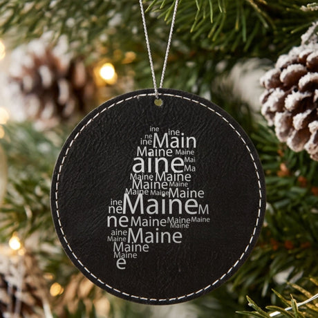 Black leatherette round Christmas ornament laser engraved with the Maine Word Cloud design, hanging on a tree