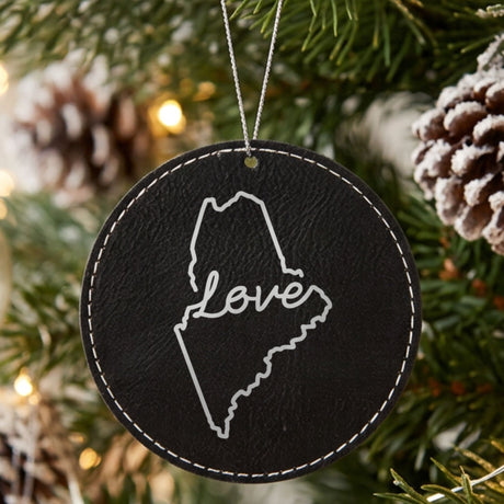 Black leatherette round Christmas ornament laser engraved with the Maine Hand-Drawn Love design, hanging on a tree