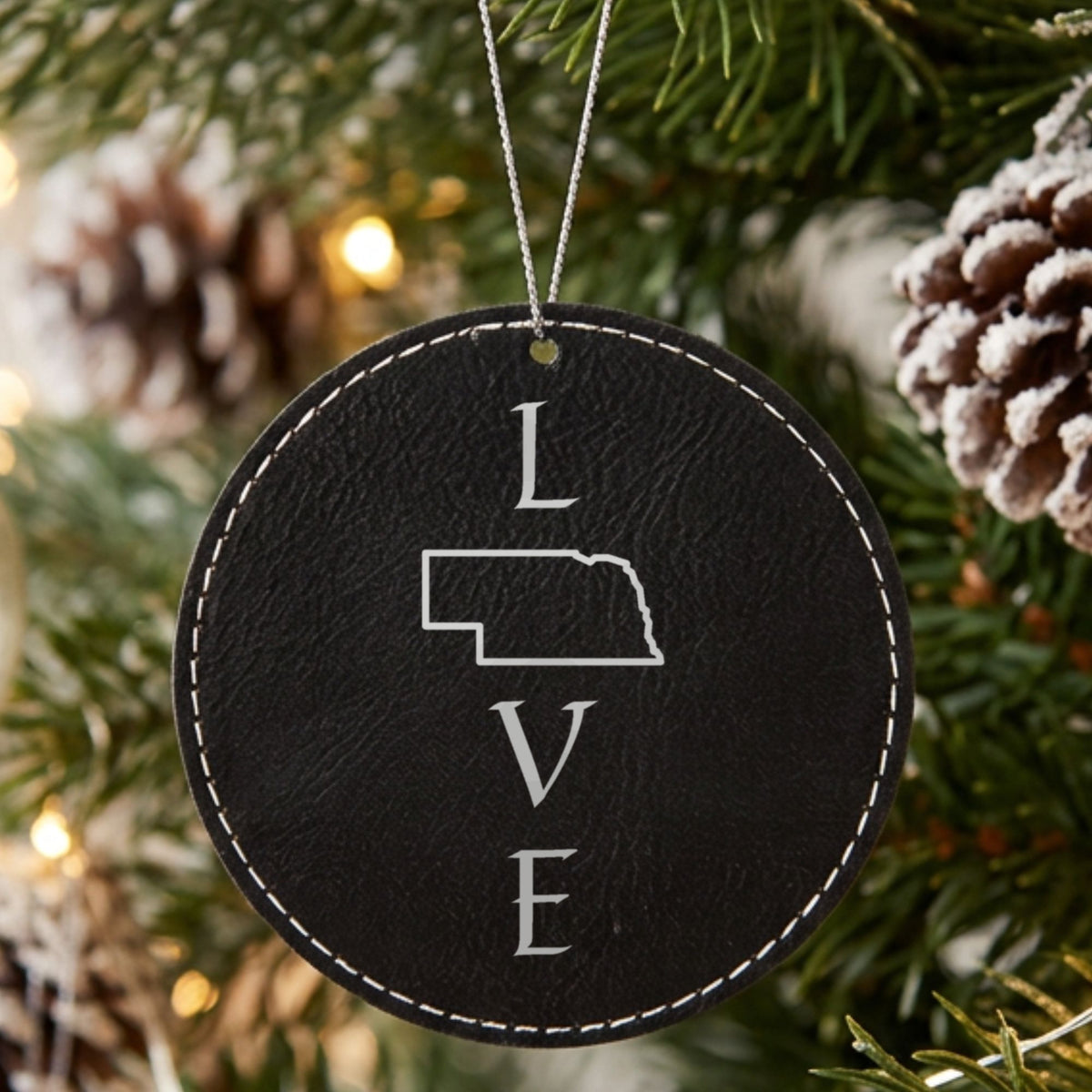 Black leatherette round Christmas ornament laser engraved with the Nebraska LOVE Stacked design, hanging on a tree
