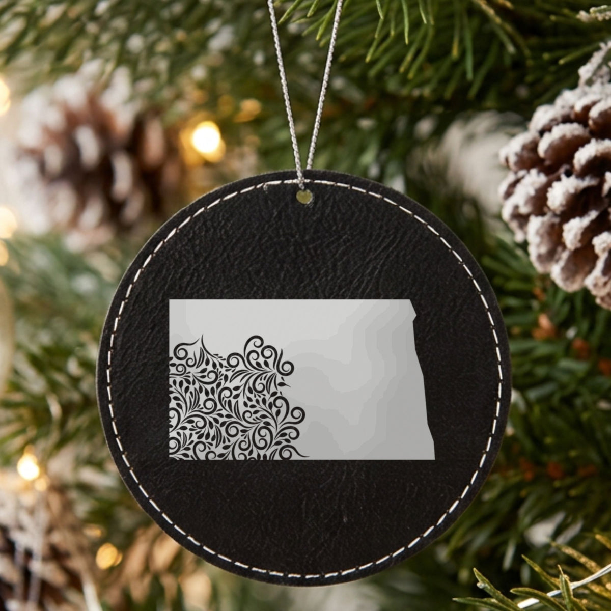 Black leatherette round Christmas ornament laser engraved with the North Dakota Floral Damask design, hanging on a tree
