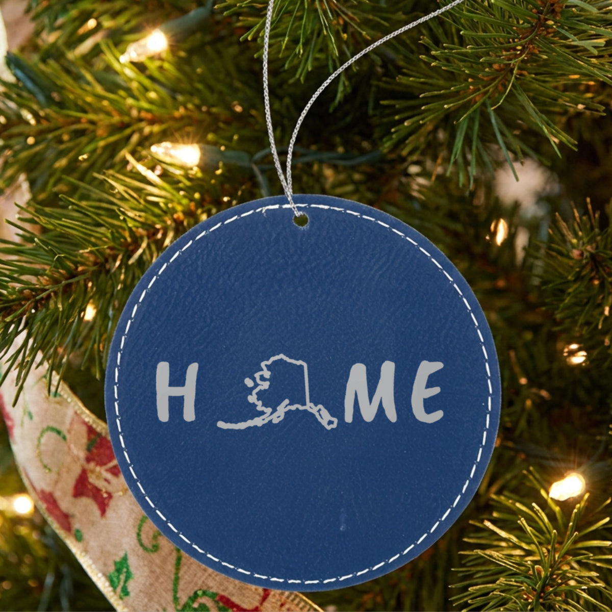 Blue leatherette round Christmas ornament laser engraved with the Alaska HOME Outline design, hanging on a tree