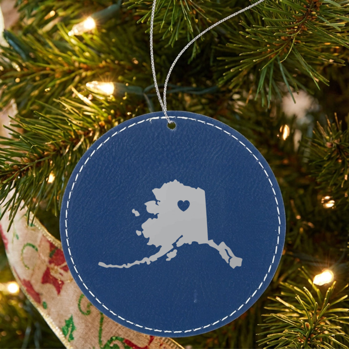Blue leatherette round Christmas ornament laser engraved with the Alaska Heart Accent design, hanging on a tree