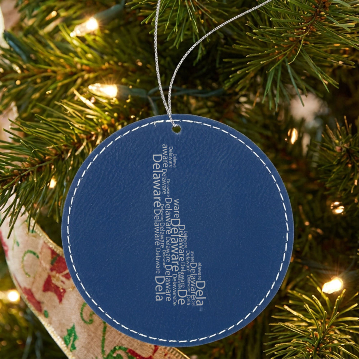 Blue leatherette round Christmas ornament laser engraved with the Delaware Word Cloud design, hanging on a tree