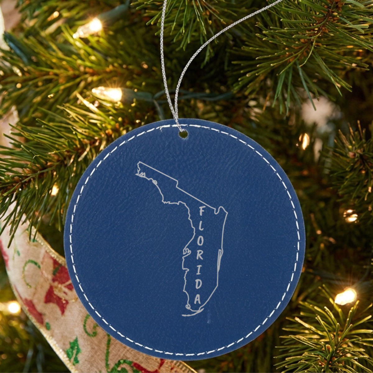 Blue leatherette round Christmas ornament laser engraved with the Florida Vertical Name (Outline) design, hanging on a tree