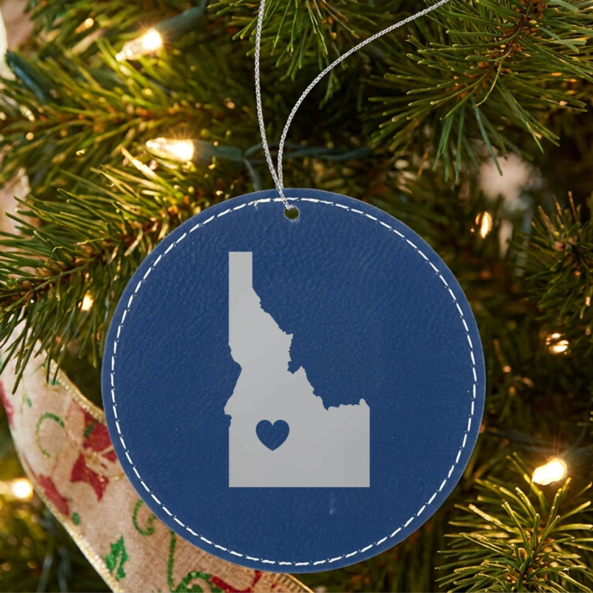 Blue leatherette round Christmas ornament laser engraved with the Idaho Heart Accent design, hanging on a tree