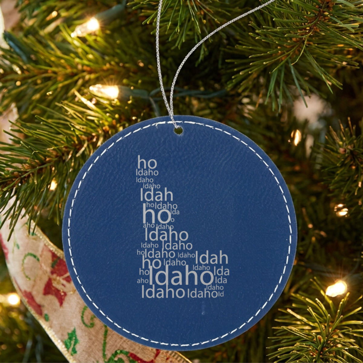 Blue leatherette round Christmas ornament laser engraved with the Idaho Word Cloud design, hanging on a tree