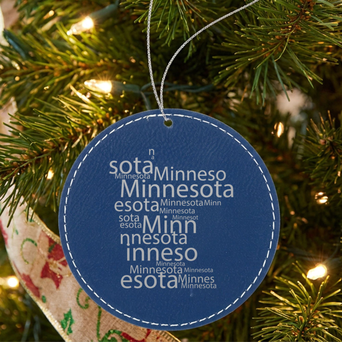Blue leatherette round Christmas ornament laser engraved with the Minnesota Word Cloud design, hanging on a tree