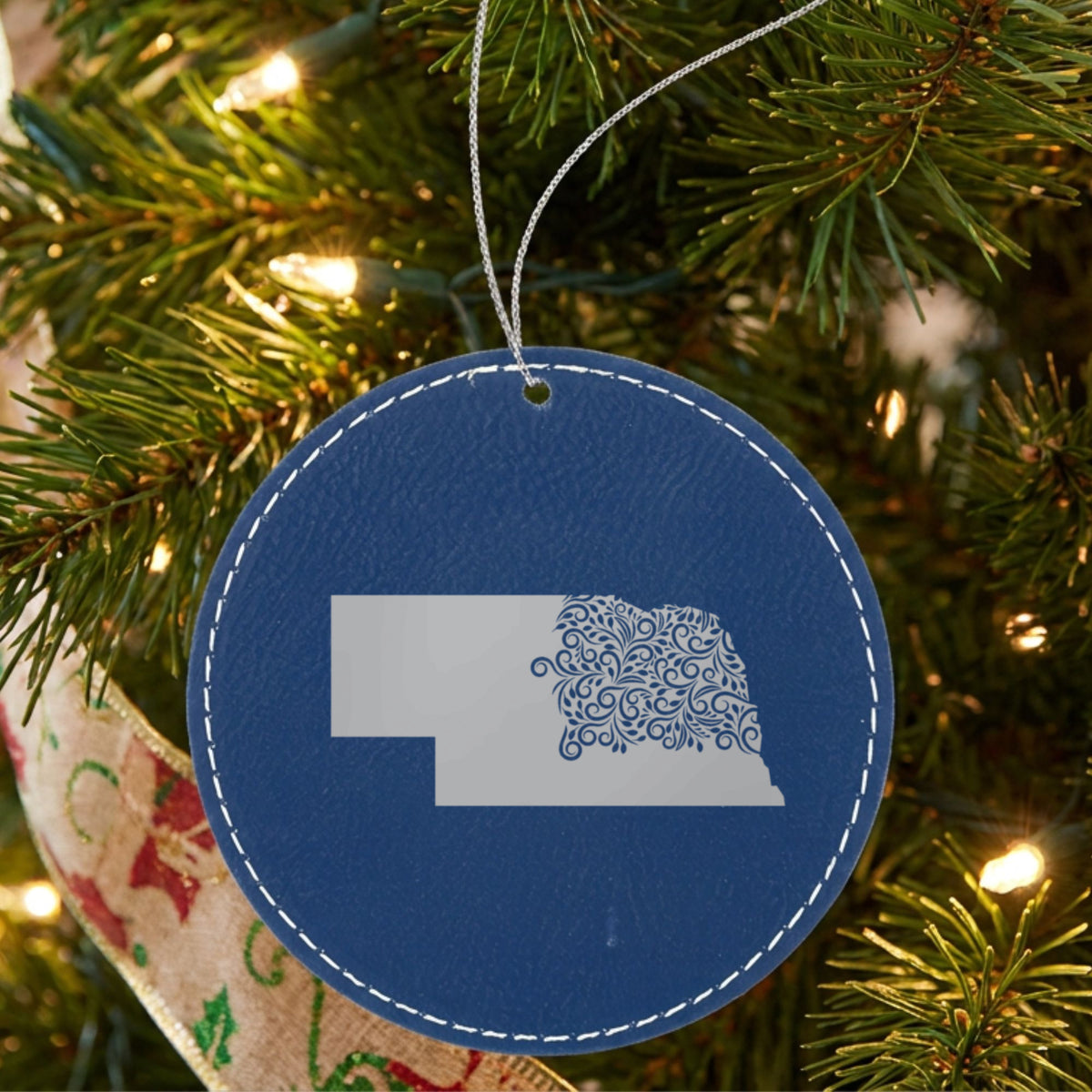 Blue leatherette round Christmas ornament laser engraved with the Nebraska Floral Damask design, hanging on a tree