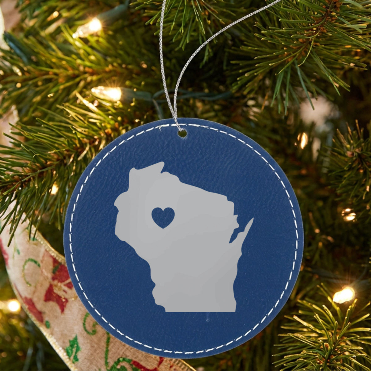 Blue leatherette round Christmas ornament laser engraved with the Wisconsin Heart Accent design, hanging on a tree