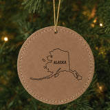 Brown leatherette round Christmas ornament laser engraved with the Alaska Brush Lettering design, hanging on a tree