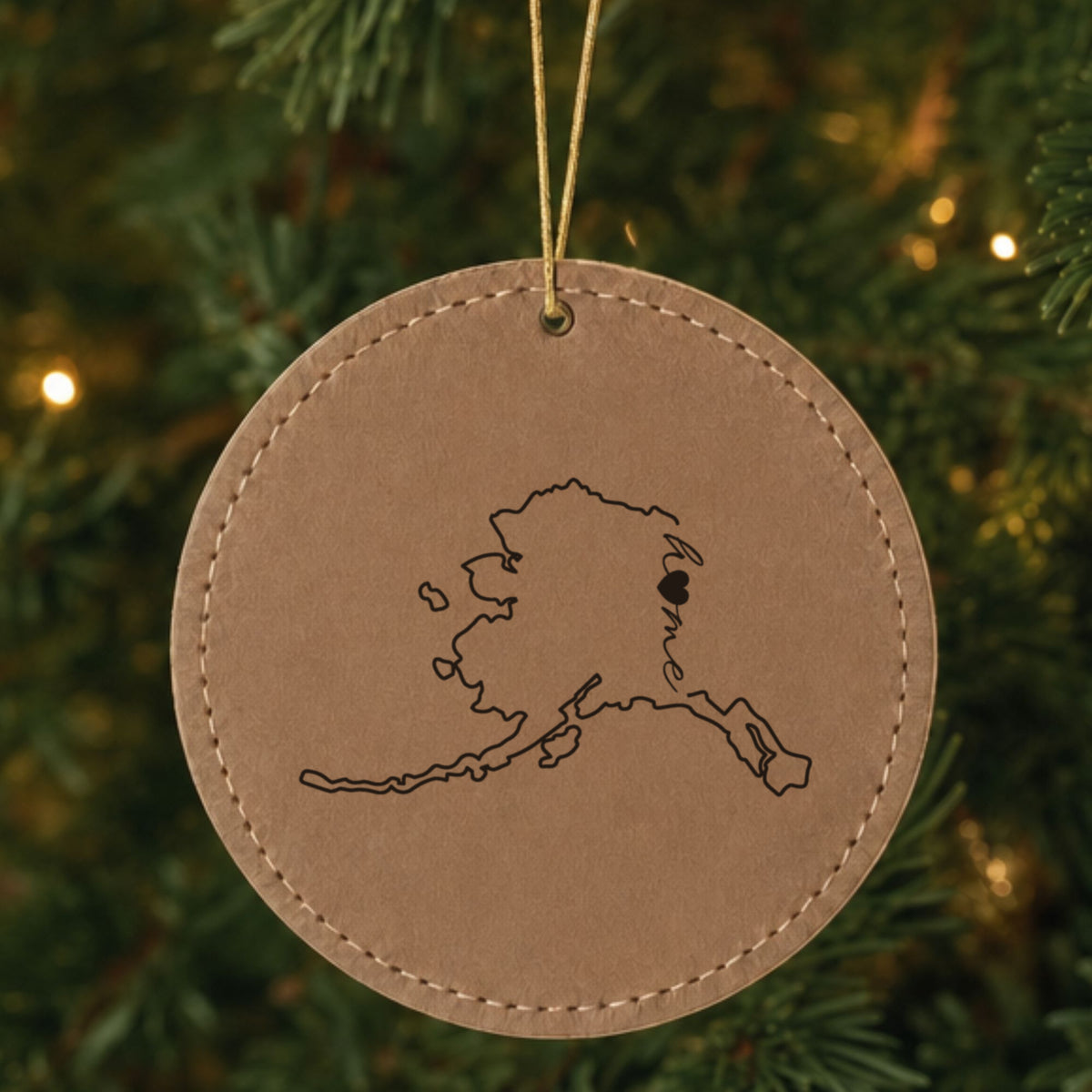 Brown leatherette round Christmas ornament laser engraved with the Alaska Home Script & Heart design, hanging on a tree