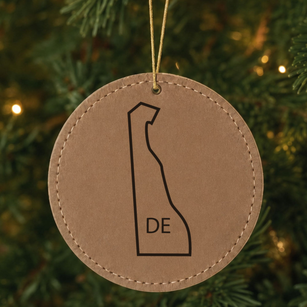 Brown leatherette round Christmas ornament laser engraved with the Delaware DE Initials design, hanging on a tree