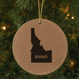 Brown leatherette round Christmas ornament laser engraved with the Idaho Bold State Name design, hanging on a tree