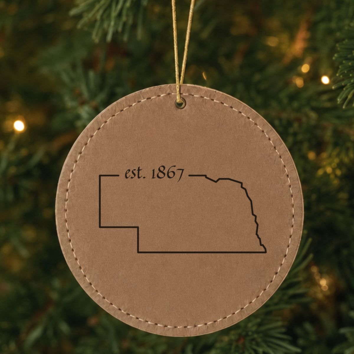 Brown leatherette round Christmas ornament laser engraved with the Nebraska Est. 1867 design, hanging on a tree