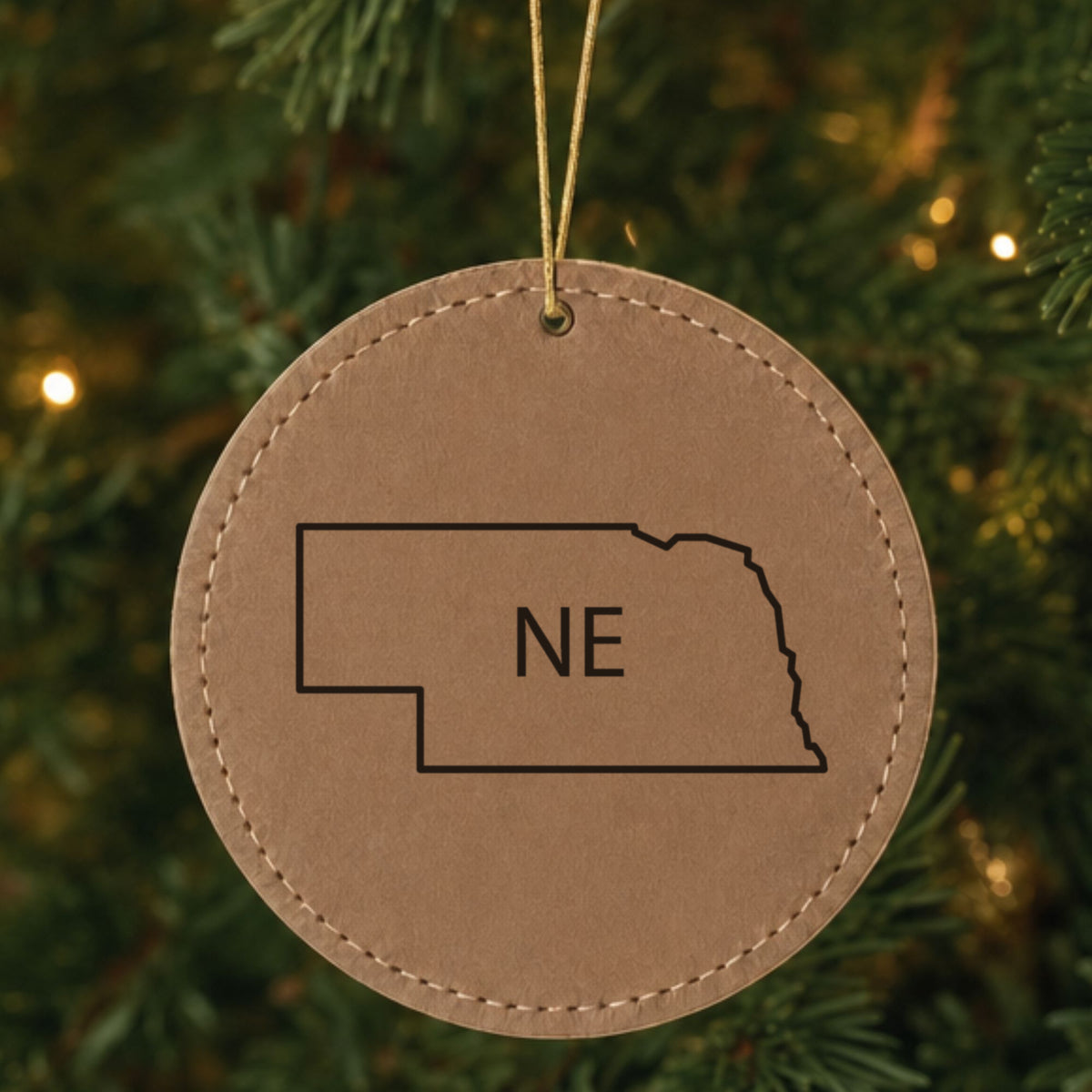 Brown leatherette round Christmas ornament laser engraved with the Nebraska NE Initials design, hanging on a tree