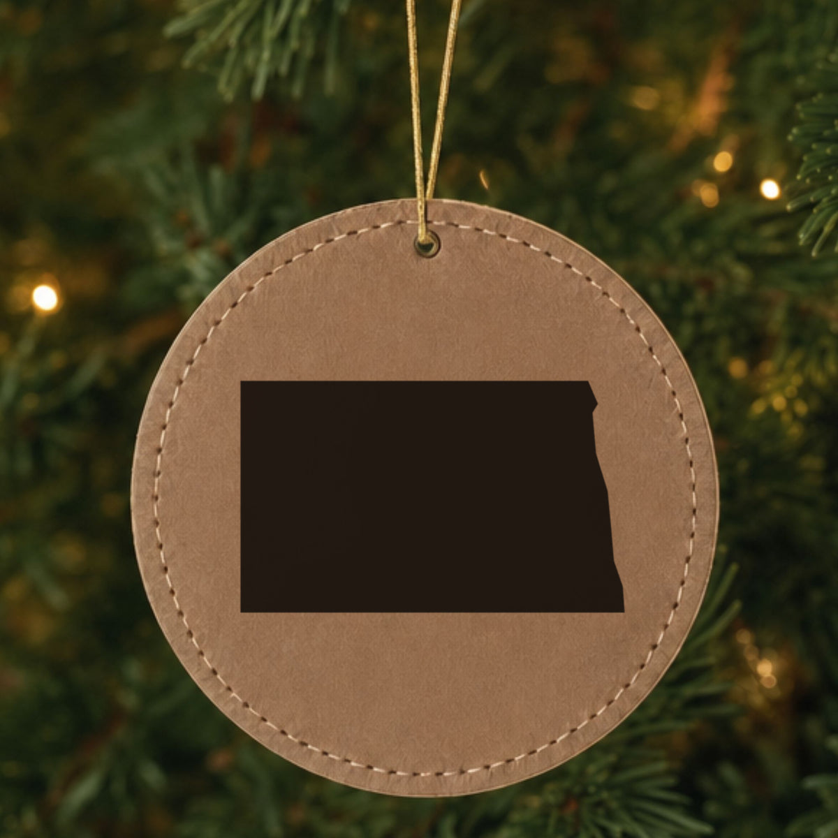 Brown leatherette round Christmas ornament laser engraved with the North Dakota Engraved State design, hanging on a tree