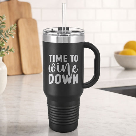 Black 40 oz tumbler with handle and straw laser engraved with "Time to Wine Down", on a kitchen counter
