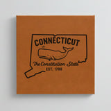 Close up of the Connecticut design engraved on Brown leatherette: a sperm whale, "The Constitution State", EST. 1788