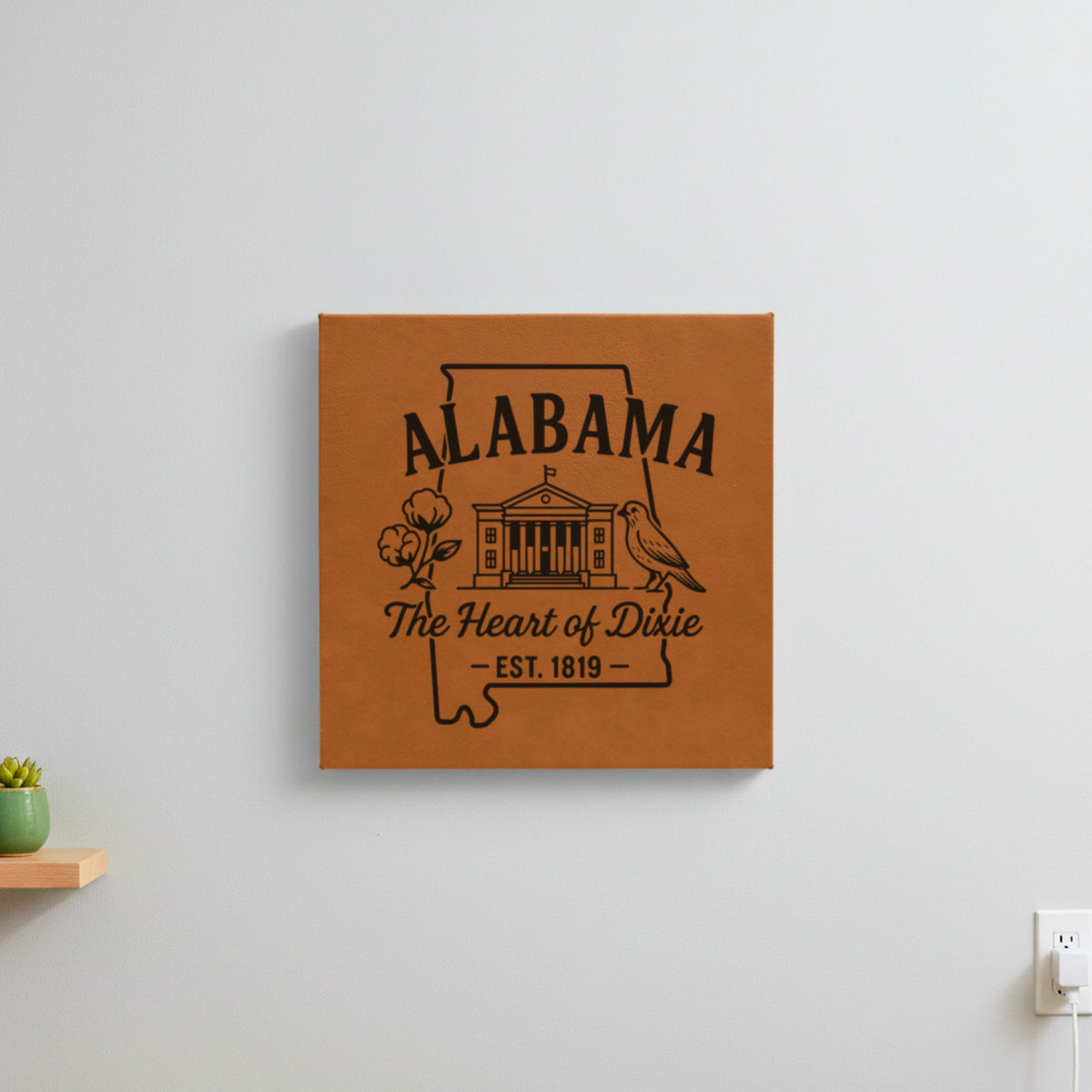 Brown leatherette wall sign engraved with an Alabama design of the capitol building, a cotton boll and a yellowhammer, hanging on a wall