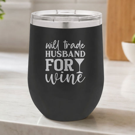 Black 12 oz stemless wine tumbler laser engraved with "Will Trade Husband for Wine", on a kitchen counter