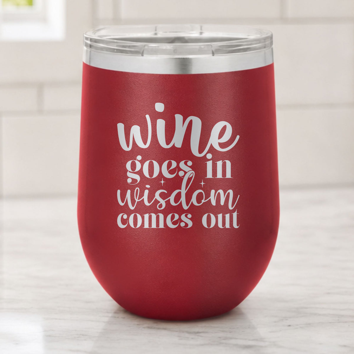Maroon 12 oz stemless wine tumbler laser engraved with "Wine Goes In, Wisdom Comes Out", on a kitchen counter